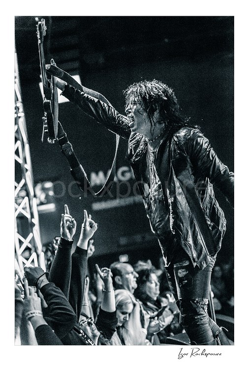Vertical black and white image of Tom Keifer performing live at the edge of the stage, raising his black guitar overhead toward the crowd in a fierce concert moment