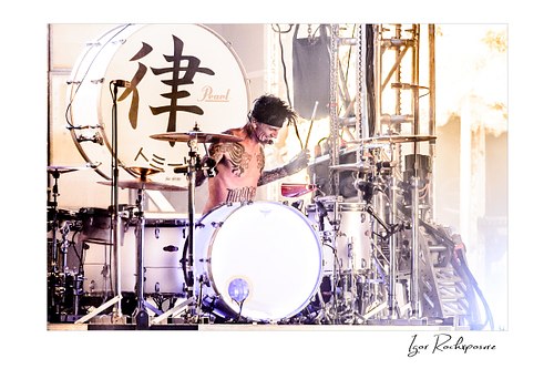 Horizontal color image of Tommy Lee performing at the drum kit during a Mötley Crüe concert, surrounded by cymbals and bright backlight