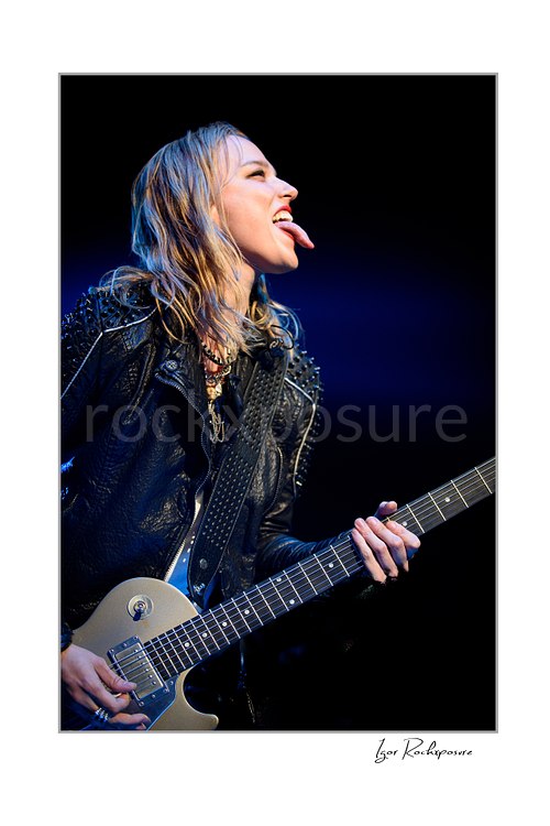 Vertical color image of Lzzy Hale of Halestorm playing guitar in a fierce live performance close-up, with tongue out, wet hair, and deep blue stage light behind her