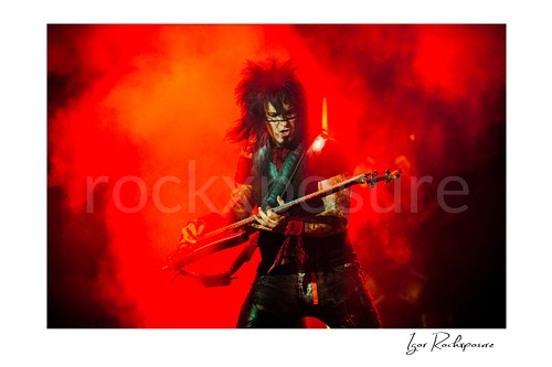 Horizontal color image of Nikki Sixx performing with bass guitar during a Mötley Crüe concert, surrounded by vivid red stage smoke