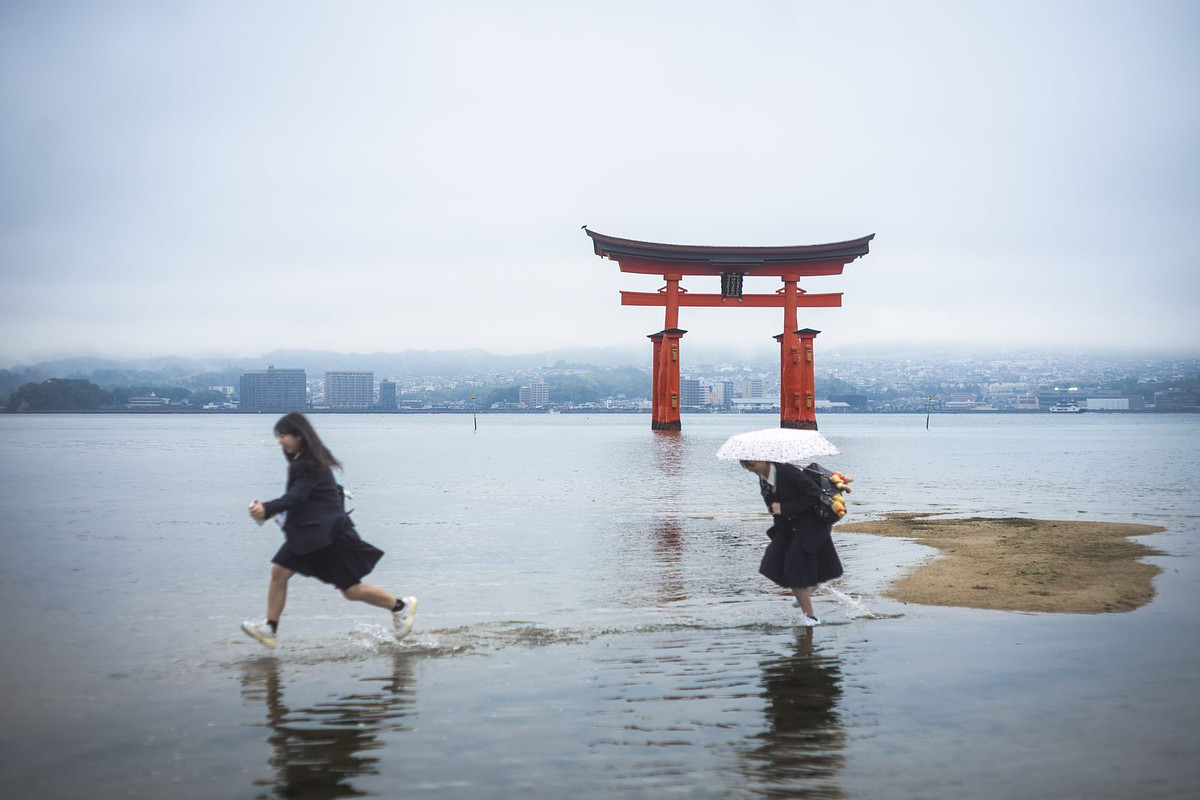 Le torii de Miyajima dans la brume, deux écolières traversent l'eau en courant, une avec un parapluie