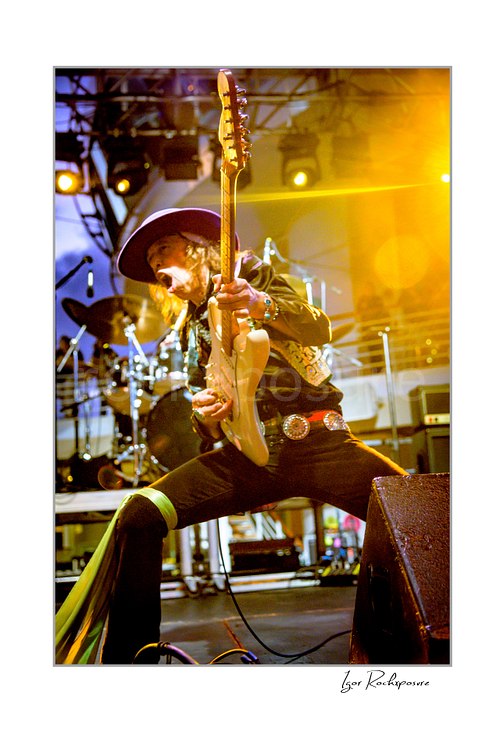 Vertical color image of Randy Hansen playing a white guitar in a wide stage stance under vivid yellow concert light on an outdoor ship deck stage