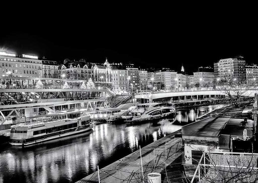 Schwarz-Weiß-Fotografie des Wiener Donaukanals bei Nacht auf Höhe Schwedenplatz mit beleuchteten Schiffen, der Schwedenbrücke und städtischer Architektur im Hintergrund.
