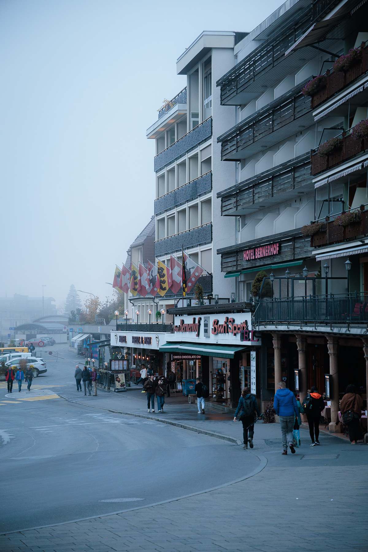 Nebelverhangene Dorfstrasse in Grindelwald mit dem beleuchteten Hotel Bernerhof, Passanten und Schweizer Flaggen an einem feuchten Herbstmorgen