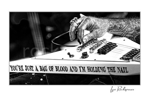 Horizontal black and white close-up image of Scott Ian’s guitar and tattooed hand on stage, showing the instrument body with the phrase “YOU’RE JUST A BAG OF BLOOD AND I’M HOLDING THE NAIL”