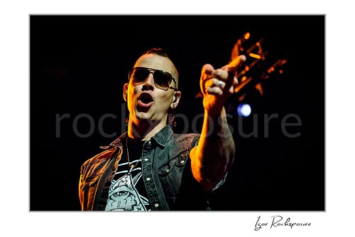 Horizontal color image of M. Shadows of Avenged Sevenfold singing onstage in sunglasses with an outstretched hand under warm concert light