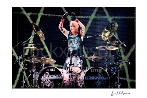 Horizontal color image of James Kottak of Scorpions behind the drums with raised arms, black gloves, and full Scorpions drum setup