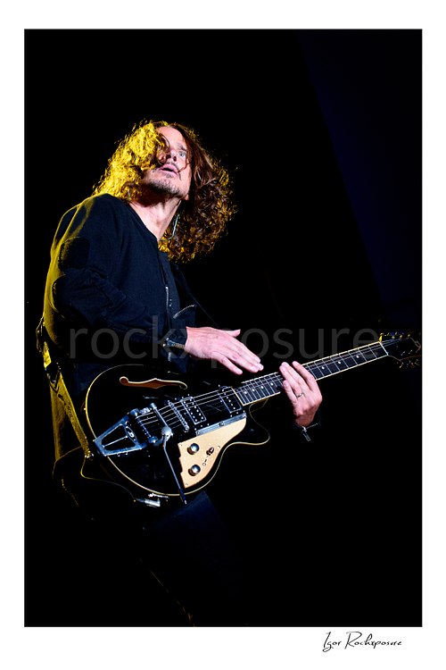 Vertical color image of Chris Cornell performing live on stage, playing electric guitar against a dark background under warm concert lighting