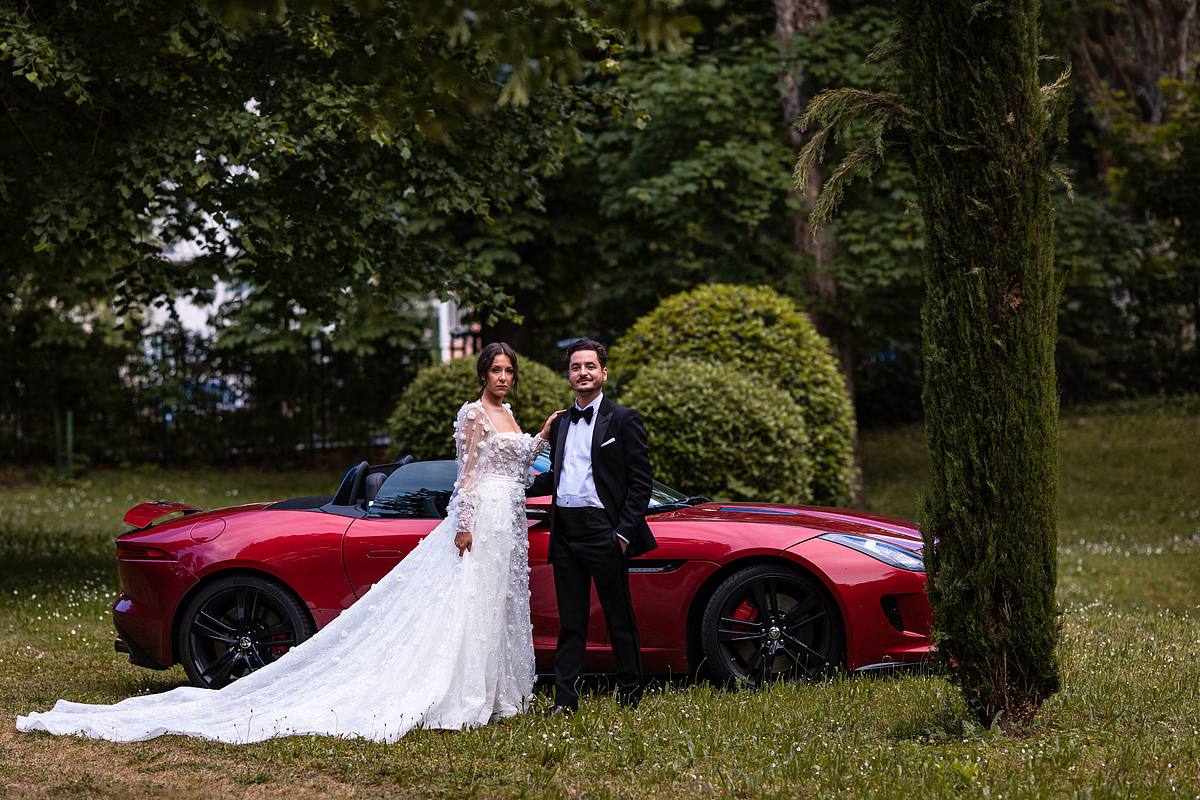 Les mari&eacute;s posent fi&egrave;rement devant une voiture de sport rouge &eacute;clatante, la mari&eacute;e &eacute;tincelante dans sa robe blanche d&eacute;taill&eacute;e, photographie saisissante par Sebastien CLAVEL Photographe mariage Lyon
