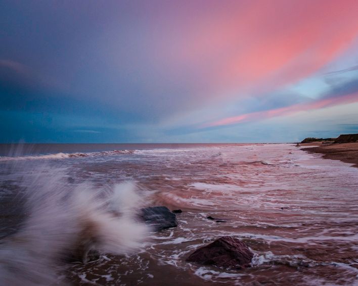Waves breaking over the rocky shore on the Norfolk coast beneath a pink and blue evening sky.