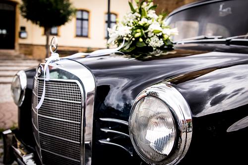 Nahaufnahme eines schwarzen Mercedes Oldtimers schräg von vorne mit glänzendem Kühlergrill, rundem Scheinwerfer und einem bunten Blumengesteck auf der Motorhaube.