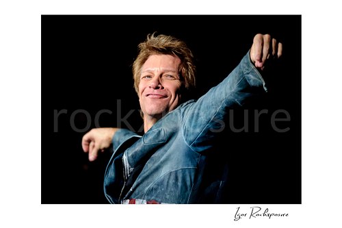 Horizontal color image of Jon Bon Jovi in a close live performance portrait, smiling with both arms raised against a dark stage background