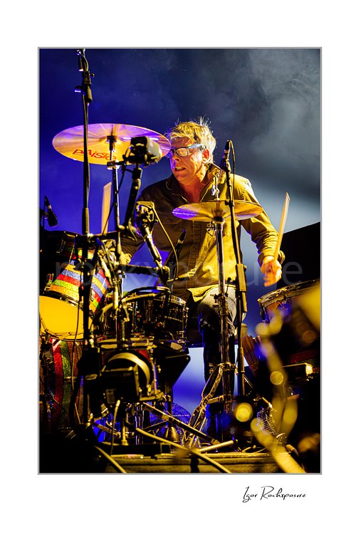 Vertical color image of Patrick Carney of The Black Keys playing drums under warm yellow and blue stage lighting with light smoke