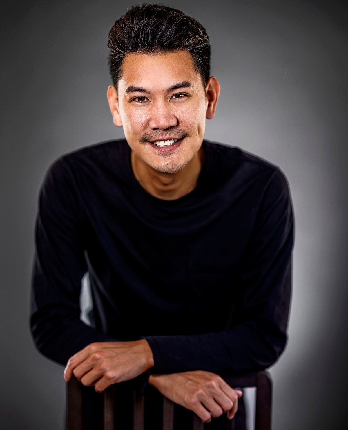 Portrait of an Asian man dressed in all black, leaning over a chair. Background graduates from light to dark on edges.