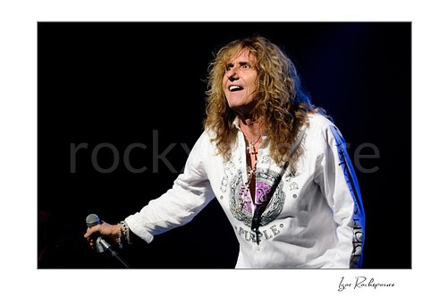 Horizontal color image of David Coverdale of Whitesnake performing live on stage, holding a microphone and mic stand under blue concert lighting