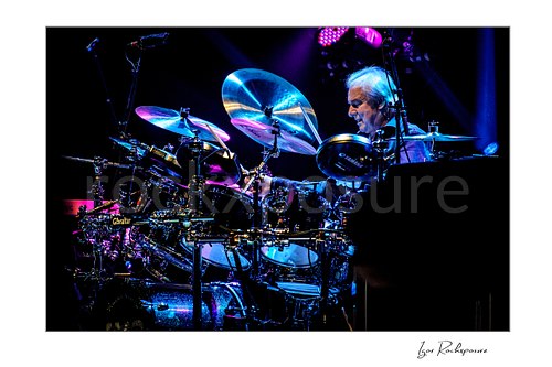 Horizontal color image of Alan White of Yes performing at the drum kit under vivid blue and purple concert lighting