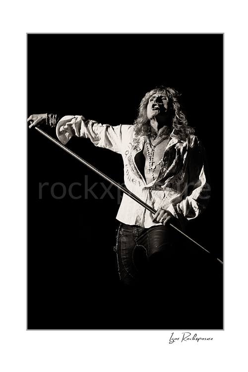 Vertical black and white image of David Coverdale of Whitesnake performing live on stage, holding a microphone stand against a dark background