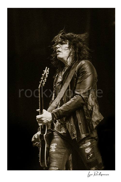 Vertical black and white image of Tom Keifer performing live on stage with a black guitar, hair blowing back in a dramatic concert portrait against a dark background