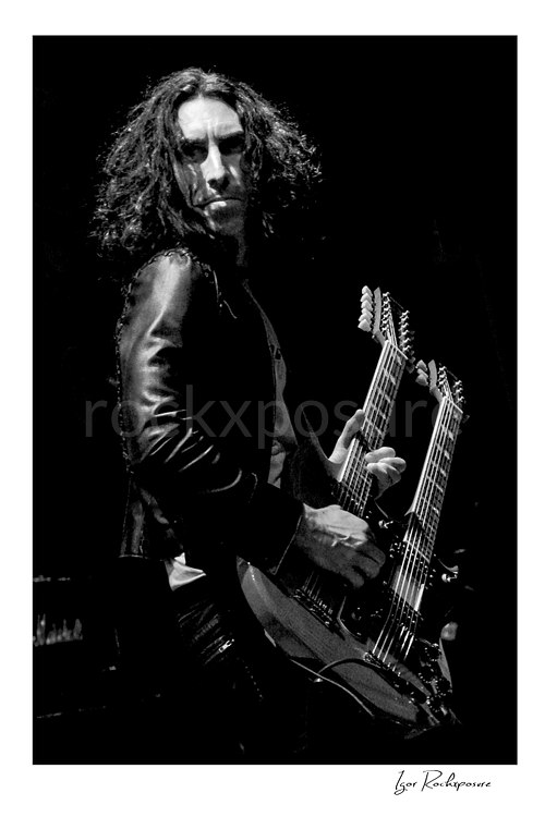 Vertical black and white image of Brent Woods holding a double-neck guitar on stage in a dramatic close-up