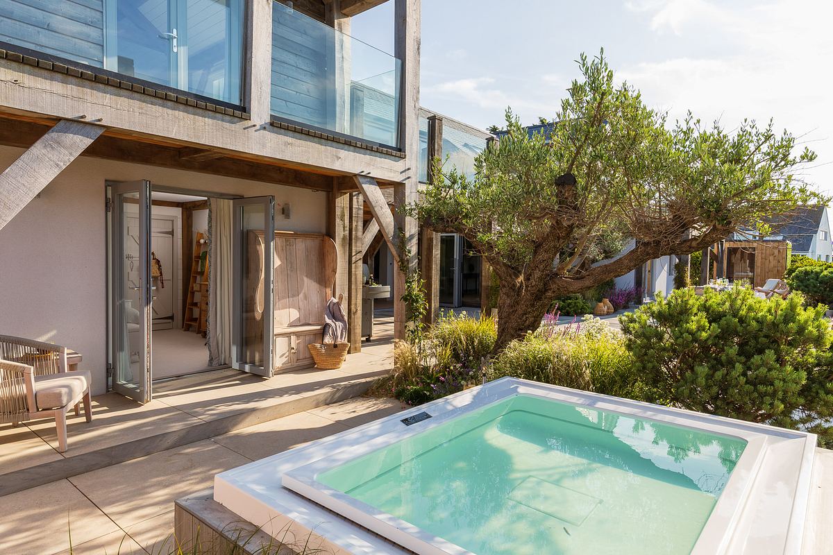 Modern outdoor patio with a hot tub, seating area, and doors leading to a bedroom; surrounded by greenery and architectural elements, expertly captured by an architecture photographer in Cornwall.