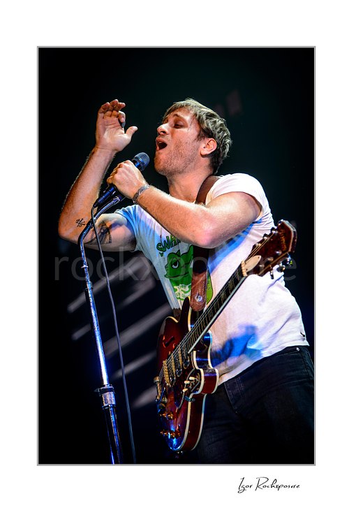 Vertical color image of Dan Auerbach of The Black Keys singing into a microphone with one hand raised while holding a guitar
