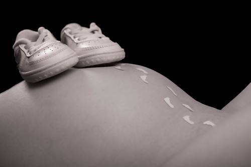 Nahaufnahme eines nackten Babybauchs in Schwarz-Weiß, auf dessen Spitze kleine weiße Babyschuhe stehen, während gemalte Fußabdrücke den Bauch hinablaufen.