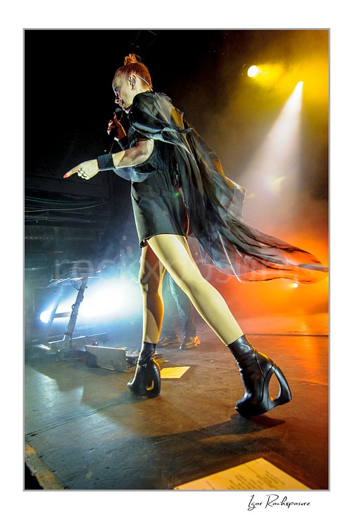 Vertical color image of Shirley Manson performing live on stage during a Garbage concert, wearing flowing black stagewear and high-heeled boots under warm concert light