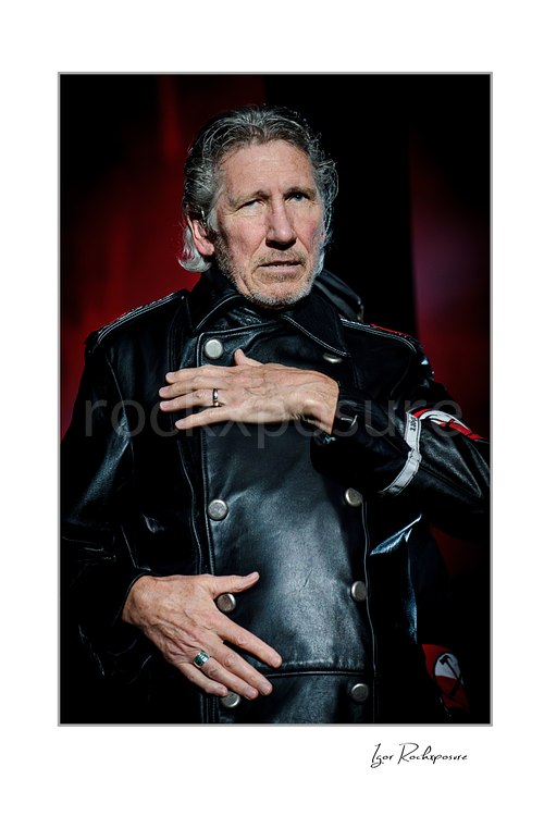 Roger Waters faces forward with one hand across his chest and another at his abdomen, wearing a black leather coat against a red background.