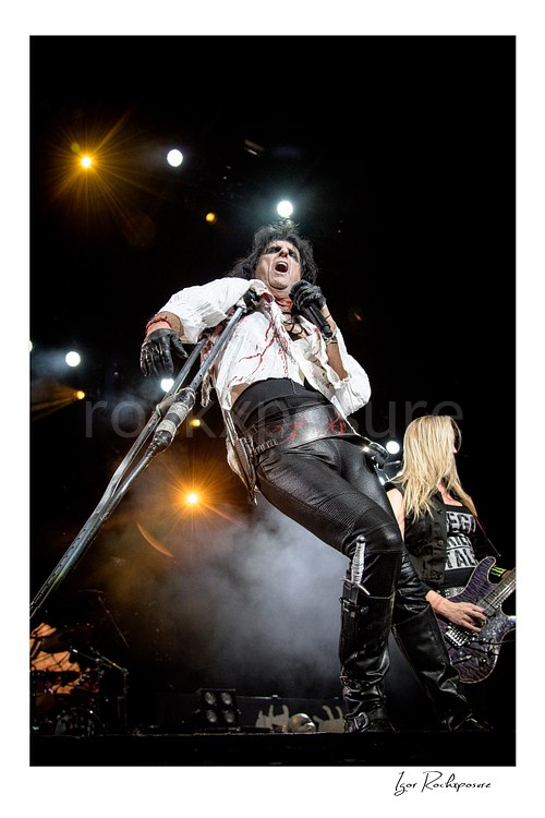 Vertical color image of Alice Cooper and Nita Strauss of the Alice Cooper Band performing live on stage with a blood-stained shirt, cane microphone stand, stage smoke, and bright overhead lights