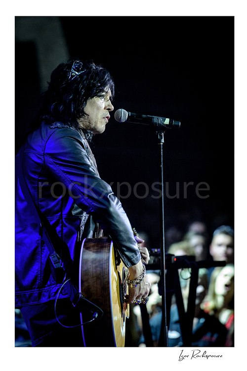 Vertical color image of Tom Keifer singing into a microphone while holding an acoustic guitar in a close live performance side profile, with blue stage light and the crowd softly blurred behind him