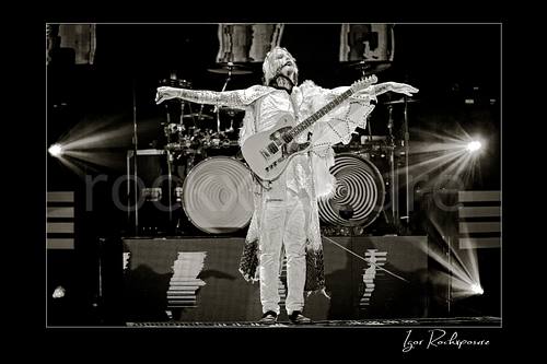 Horizontal black and white image of John 5 performing live on stage with an electric guitar, white stage outfit, outstretched arms, and bright backlights