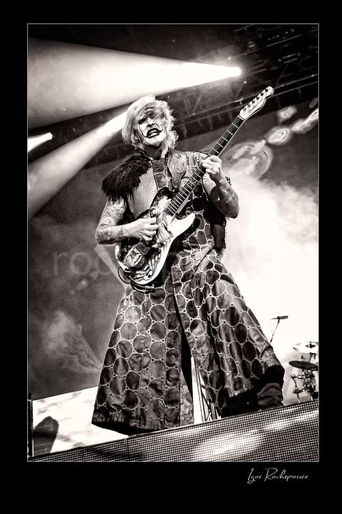Vertical black and white image of John 5 performing live on stage with an electric guitar, theatrical makeup, patterned stage outfit, and bright backlighting