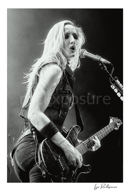 Vertical black and white image of Nita Strauss performing live on stage with an electric guitar beside a microphone