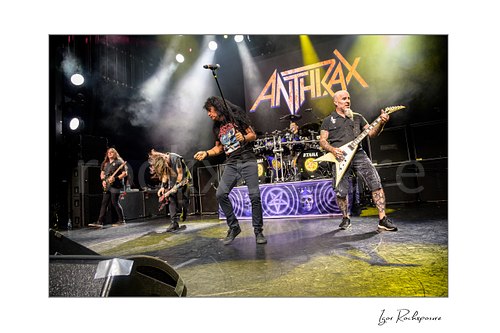 Horizontal color image of Frank Bello, Jonathan Donais, Joey Belladonna, Charlie Benante, and Scott Ian of Anthrax performing live on stage, with the full band visible beneath the Anthrax logo under bright concert lighting