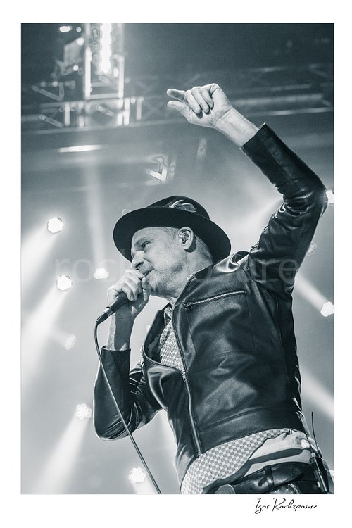 Vertical black and white image of Gord Downie of The Tragically Hip singing into a microphone with one arm raised, wearing a hat and leather jacket under bright stage lights