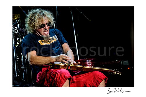 Horizontal color image of Sammy Hagar seated onstage playing lap steel guitar in sunglasses and red shorts during a live performance