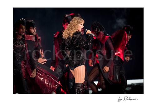 Horizontal color image of Taylor Swift singing on stage in profile with dancers and red concert lighting