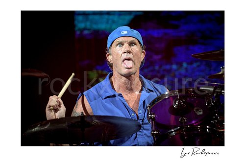 Chad Smith of Red Hot Chili Peppers sticks out his tongue with eyes wide, holding a drumstick above cymbals beside a purple tom and colorful LEDs.