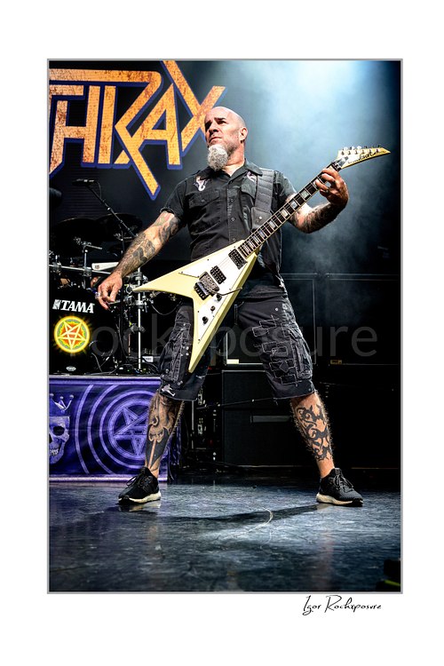 Vertical color image of Scott Ian of Anthrax performing live on stage, playing guitar in a wide stance under concert lighting