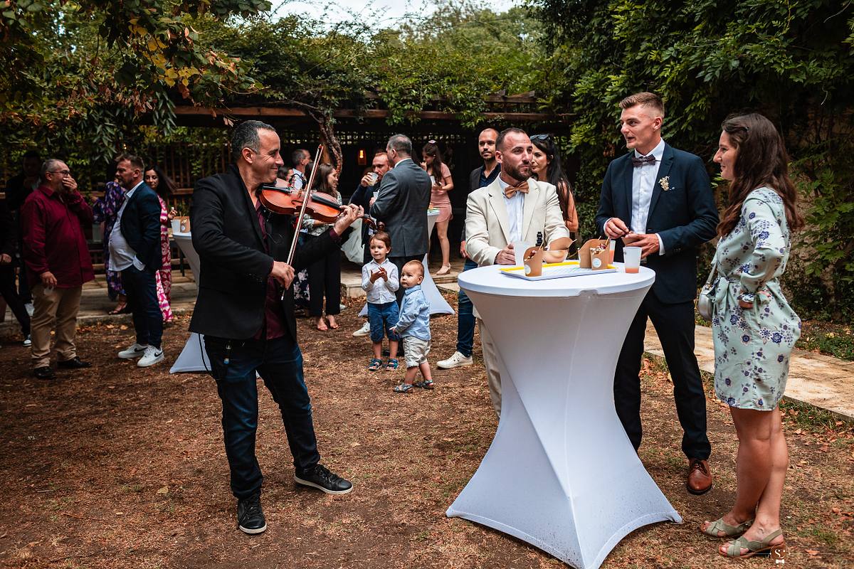 Convivialit&eacute; post-c&eacute;r&eacute;monie avec un violoniste ambulant, captant l'attention des invit&eacute;s, une atmosph&egrave;re festive r&eacute;v&eacute;l&eacute;e par S&eacute;bastien Clavel, photographe de mariage actif dans toute la France