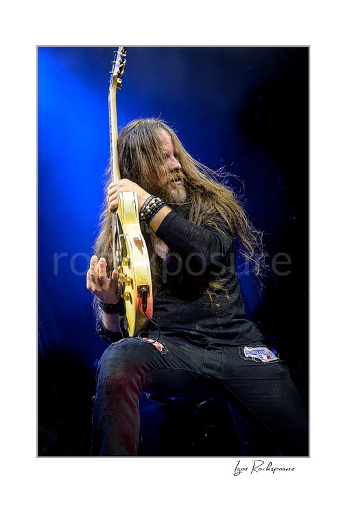 Vertical color image of Chris Latham of Beasto Blanco playing guitar under vivid blue stage light, with long hair falling across his face in a dramatic live performance pose