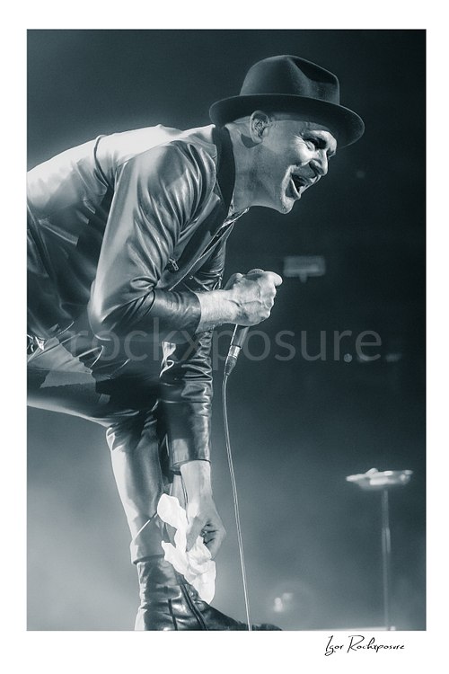 Vertical black and white image of Gord Downie of The Tragically Hip leaning forward and singing into a microphone, wearing a hat and leather jacket with a white cloth in hand amid bright concert haze