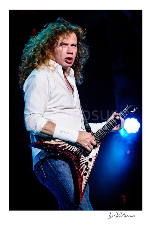 Vertical color image of Dave Mustaine of Megadeth playing guitar on stage with curly hair and bright blue stage light