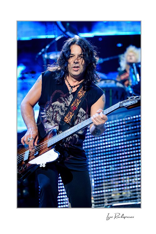 Vertical color image of Paweł Mąciwoda of Scorpions playing bass live with long dark hair and bright blue stage lights