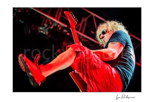 Horizontal color image of Sammy Hagar playing guitar onstage in sunglasses with a high kick under vivid red concert lighting