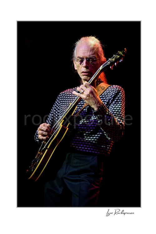 Vertical color image of Steve Howe of Yes playing guitar onstage under warm red concert light