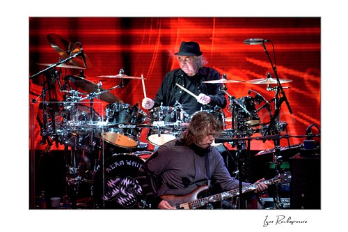 Horizontal color image of Alan White and Billy Sherwood of Yes performing onstage together with a vivid red concert backdrop
