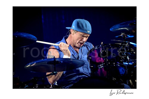 Chad Smith looks down at his purple drum kit with a drumstick raised beside his shoulder, surrounded by cymbals under deep blue stage light.