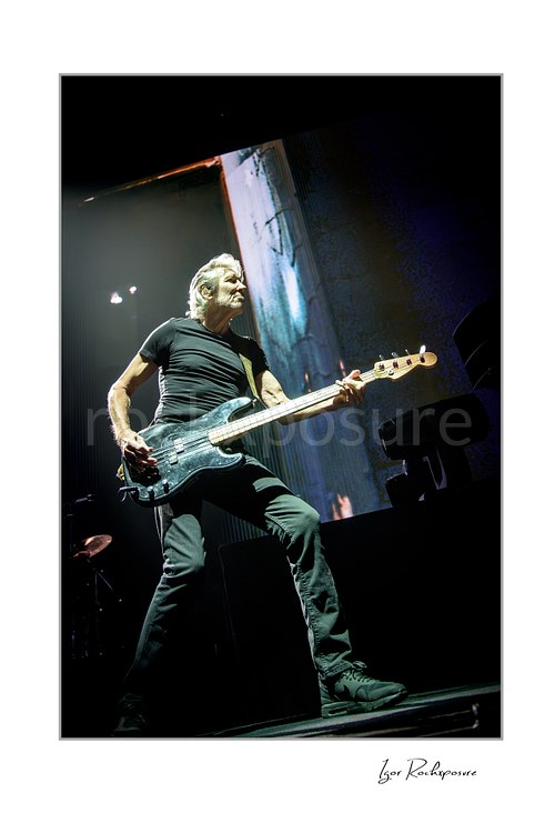 Roger Waters plays black bass in a wide stance at the stage edge, seen from below with a tall projected screen behind him.
