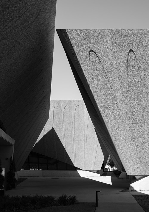 Low-angle fine art black and white photograph capturing the dramatic structural tension where the outwardly canting walls of Sir David Adjaye's Winter Park pavilions lean toward one another against the sky.
